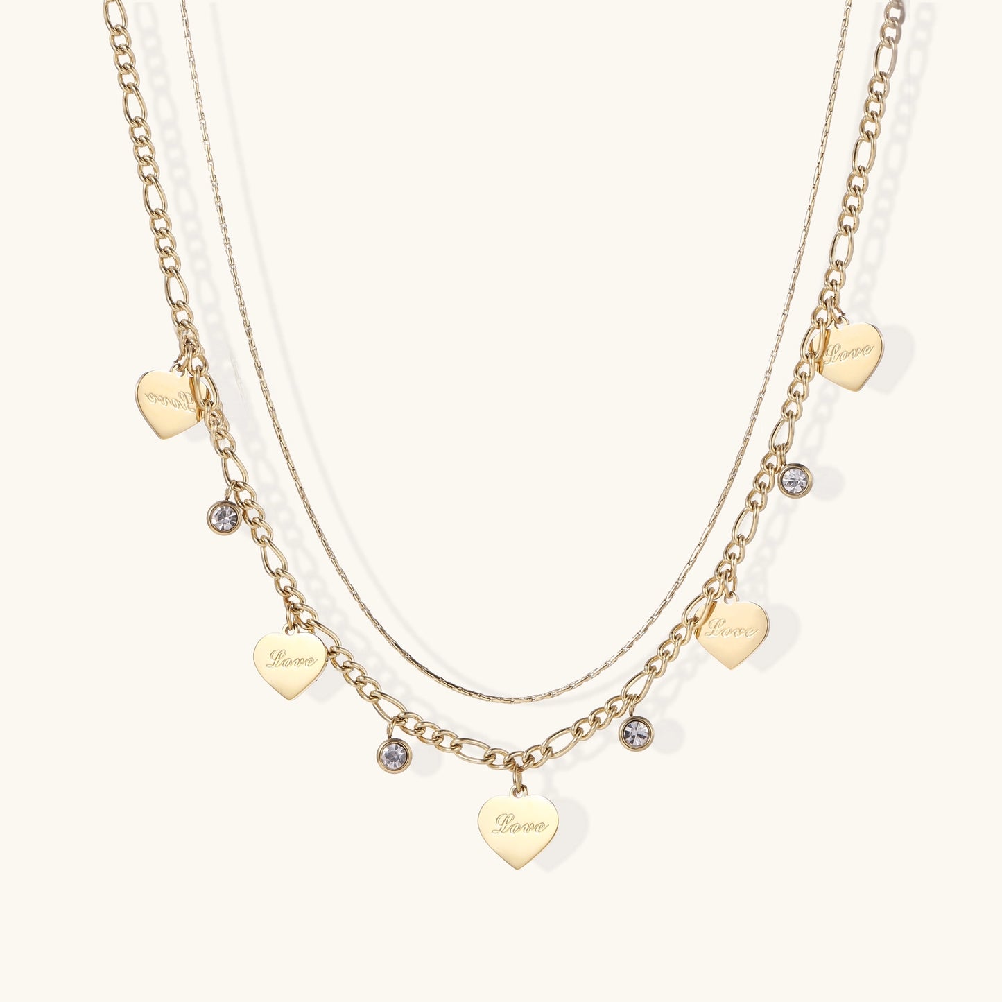 Two layered gold-plated necklaces with heart pendants engraved 'Love' and sparkling crystal charms on white background.